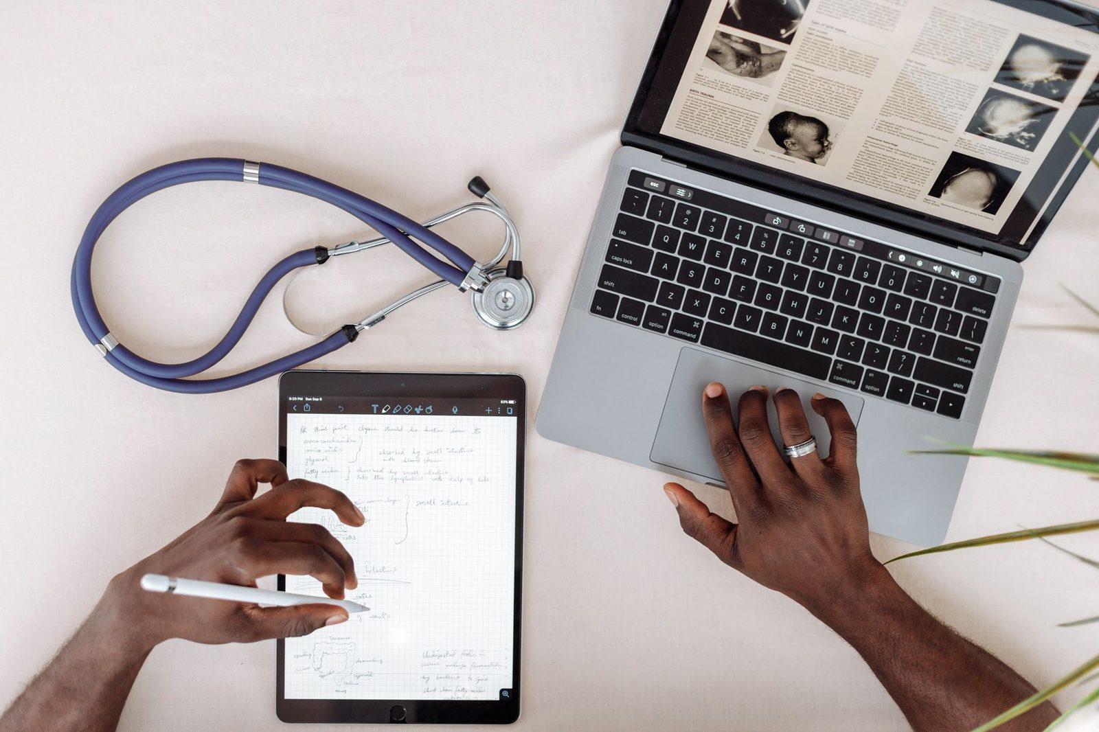 Healthcare professional reviewing information on a tablet and laptop with a stethoscope on the desk